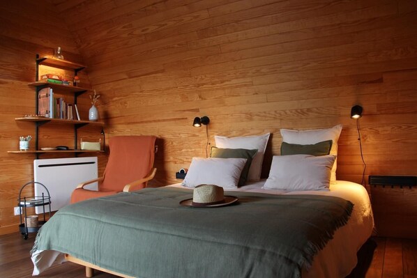 A cozy bedroom with a wooden ceiling, a bed with multiple pillows, a chair, a desk, and shelves with books and decorative items.