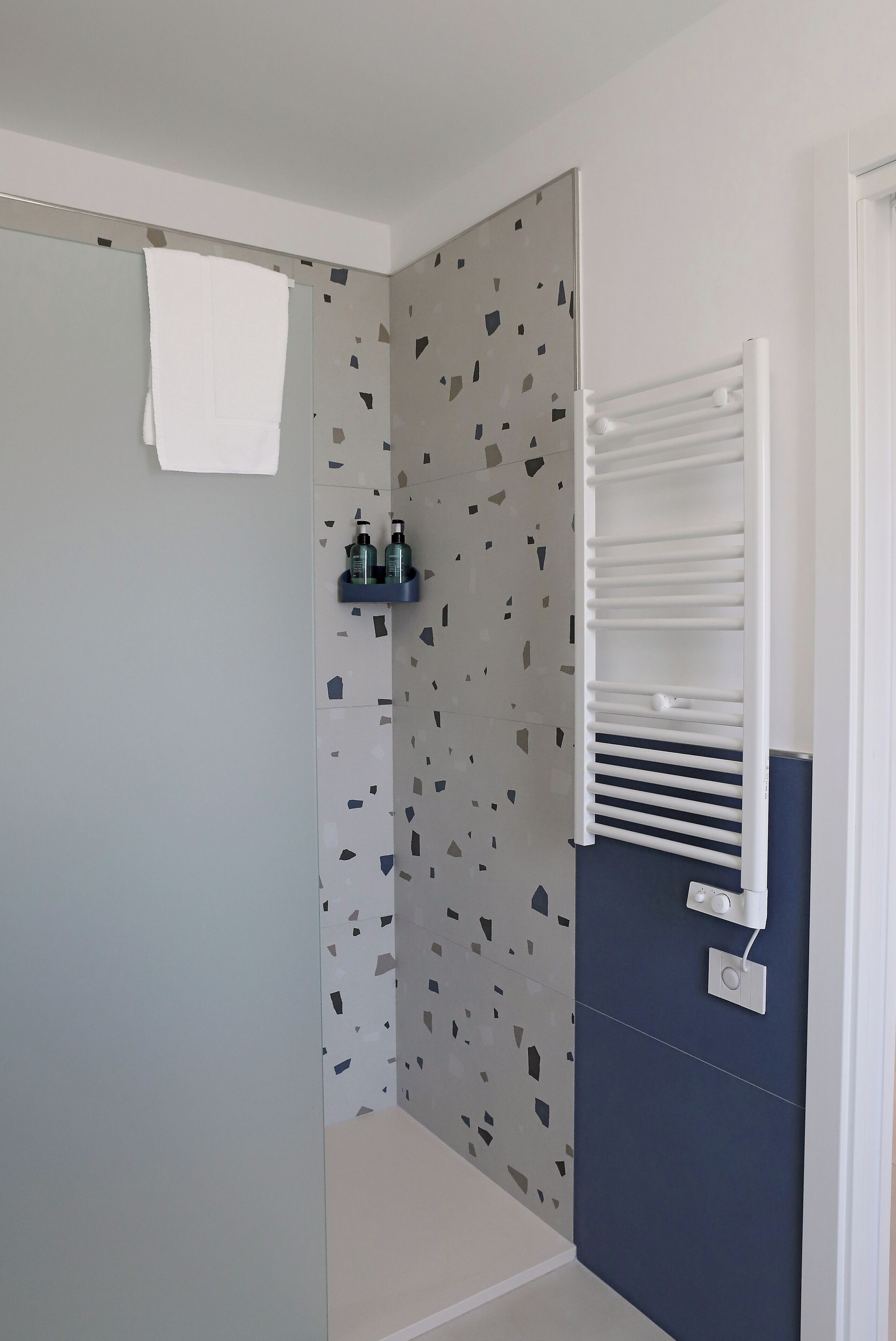 A bathroom with a shower, a towel hanging on the door, a shelf with toiletries, and a towel rack.