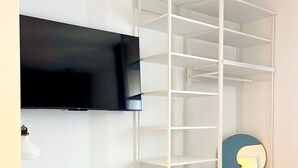 Ein Zimmer mit Schreibtisch, Stuhl, Fernseher und einem weißen Regal.