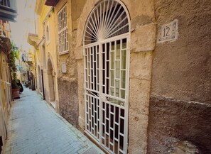 Una calle estrecha flanqueada por edificios, con un sendero pavimentado y un amplio ventanal de arco con rejas a la derecha.