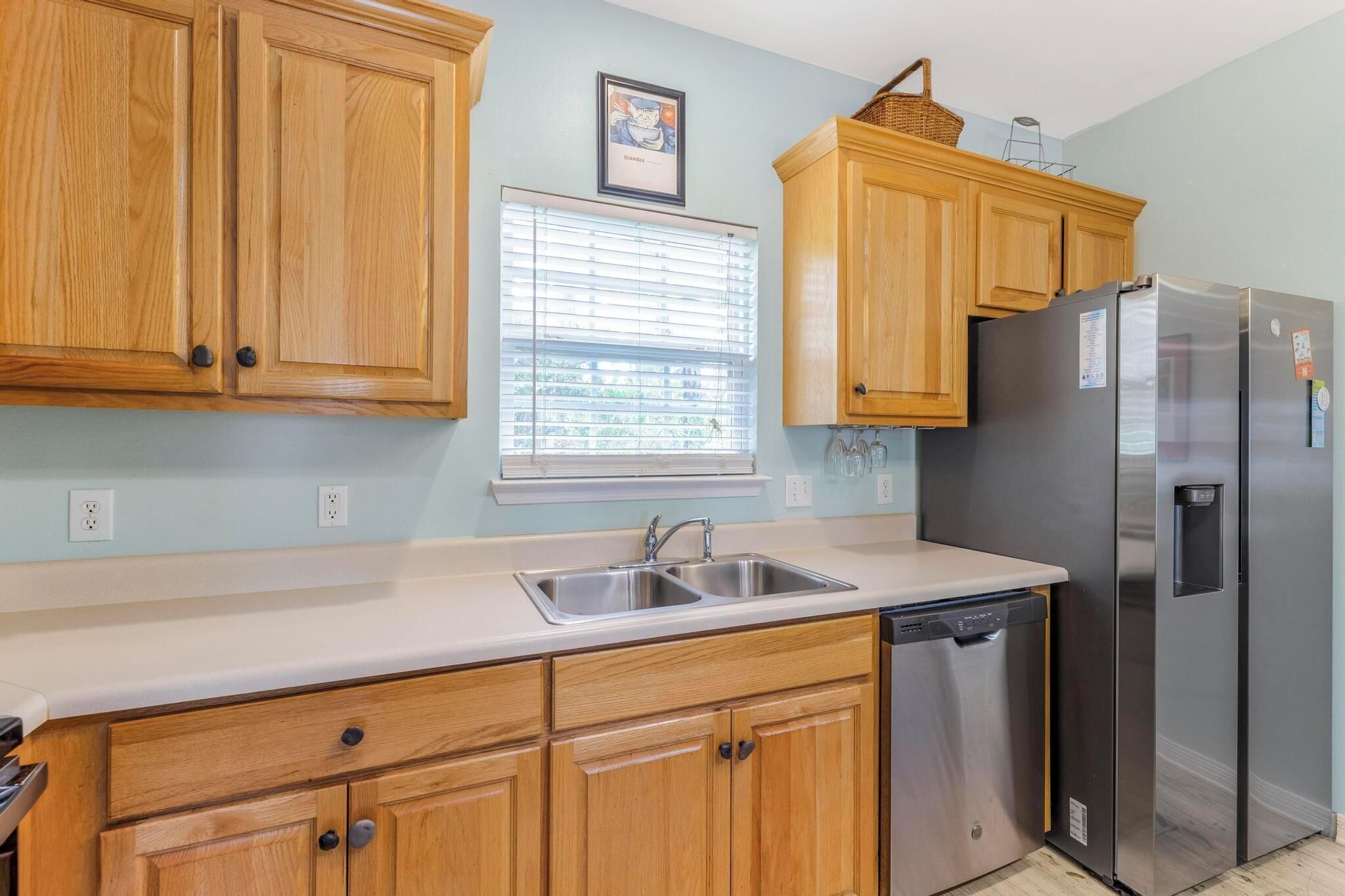 A kitchen with wooden cabinets, a stainless steel refrigerator, a dishwasher, and a window with blinds.