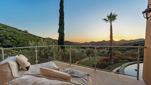 A balcony with a sofa and cushions, overlooking a landscape with trees and hills.