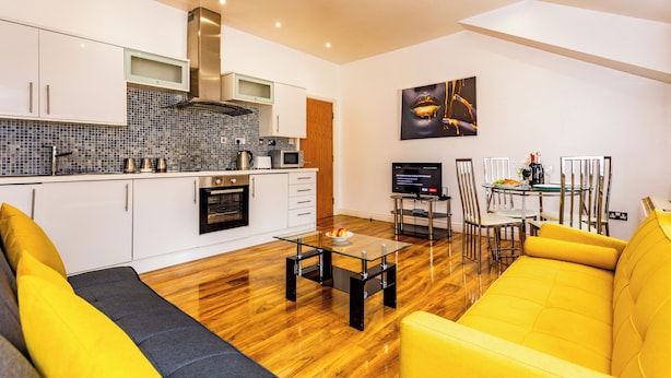Una cocina moderna con armarios blancos, un microondas y una estufa. Hay un comedor con mesa y sillas de vidrio, y un sofá amarillo en la sala de estar.