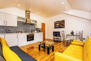 Una cocina moderna con armarios blancos, un microondas y una estufa. Hay un comedor con mesa y sillas de vidrio, y un sofá amarillo en la sala de estar.