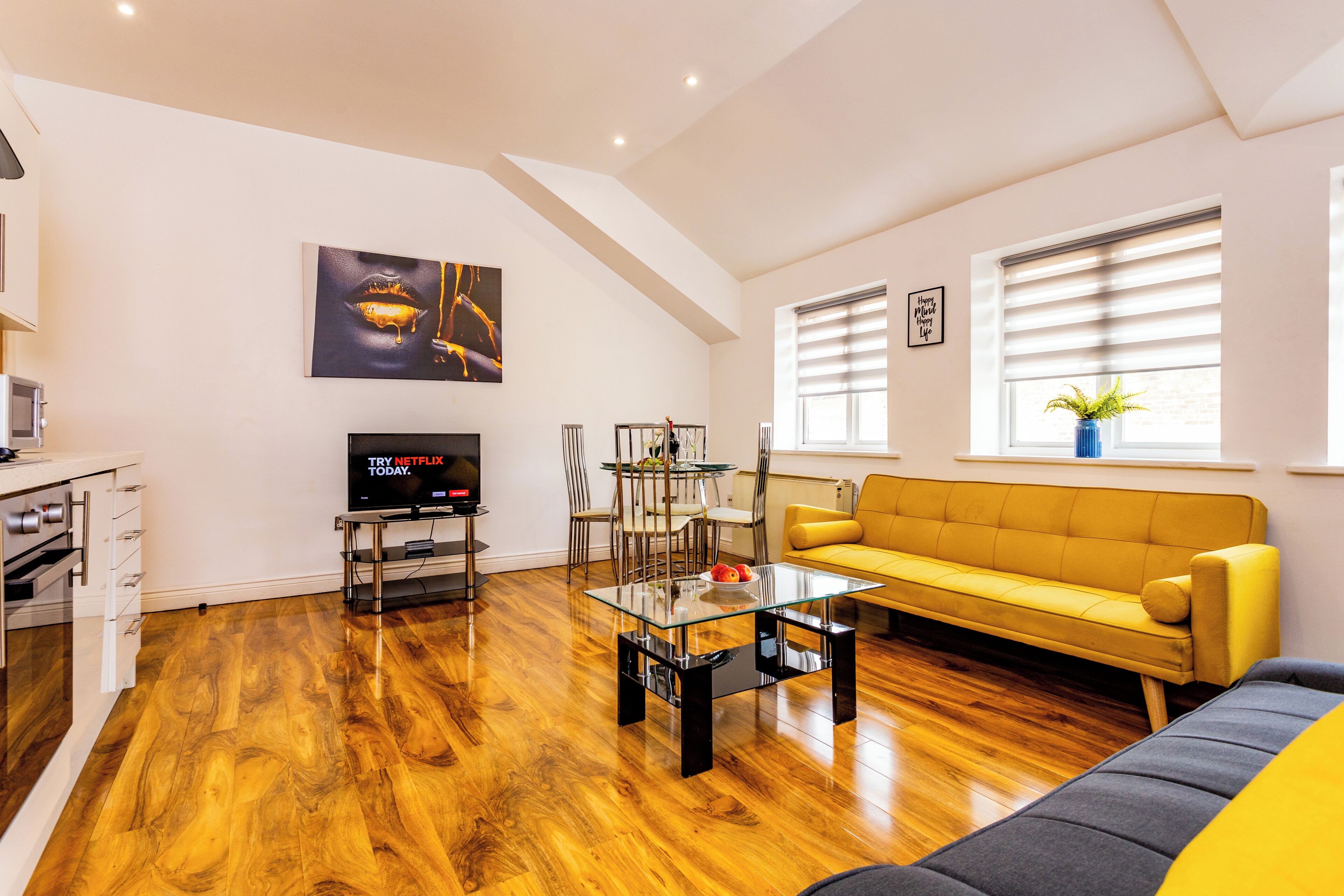Un salon moderne avec un plancher en bois, un canapé jaune, une table basse en verre, un téléviseur et un coin repas comprenant des chaises et une table.