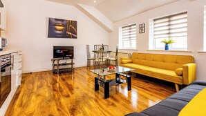 Una sala moderna con piso de madera, un sofá amarillo, una mesa de centro de vidrio, un televisor y un comedor con sillas y una mesa.