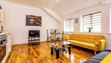 Un salon moderne avec un plancher en bois, un canapé jaune, une table basse en verre, un téléviseur et un coin repas comprenant des chaises et une table.