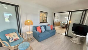 Een woonkamer met een blauwe bank, een rieten fauteuil, een kleine ronde tafel, een lamp en een tv op een meubel.