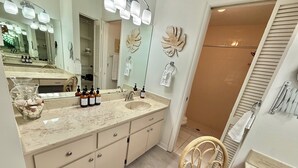 Een badkamer met een grote spiegel, een wastafelmeubel met een wastafel, diverse flesjes shampoo en een douchegedeelte.