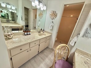 A bathroom with a large mirror, vanity with a sink, multiple bottles of shampoo, and a shower area. - Sea Breeze - a perfect place for your vacation. (Port Charlotte)