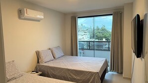 Een hotelkamer met twee bedden, een plafondlamp, een televisie en uitzicht op gebouwen en bomen.