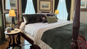 Una cama con dosel, con una colcha acolchada, dos lámparas de noche, una mesita redonda con un florero y cuadros enmarcados en las paredes.
