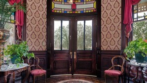 Una habitación con una puerta de madera grande, papel tapiz estampado, dos sillas, una alfombra redonda y una planta en maceta.