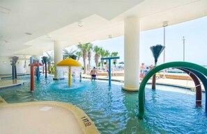 Et indendørs poolområde med vandrutsjebaner, palmer og et overdækket loungeområde.