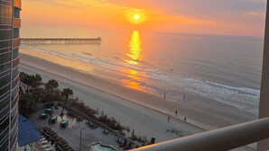 Une vue sur le coucher de soleil depuis un balcon donnant sur une plage, avec une jetée, une piscine et des chaises longues.