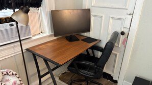 Habitació amb escriptori de fusta, cadira d'oficina negra, pantalla, làmpada de peu i radiador.