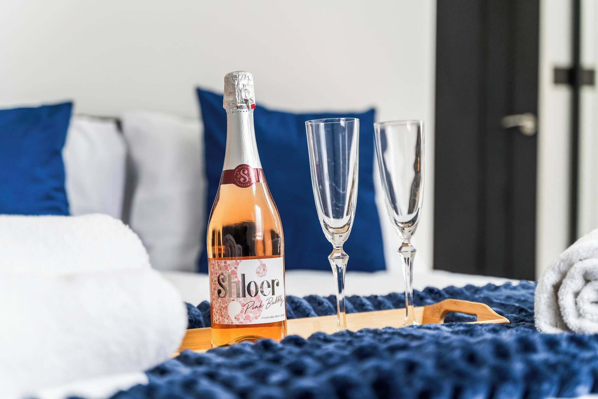 A bottle of Shloer Pink Bubble on a tray, two glasses, and a blue blanket on a bed with white and blue pillows.