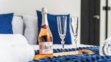 A bottle of Shloer Pink Bubble on a tray, two glasses, and a blue blanket on a bed with white and blue pillows.