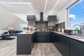 A modern kitchen with a central island, black cabinetry, and a large window providing a view. - Luxury 2 Bed Penthouse With Views Over London With Free Parking (Uxbridge)
