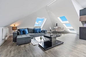 A modern loft apartment with a sloped ceiling, skylights, a grey sofa, a glass coffee table, and a small dining area. - Luxury 2 Bed Penthouse With Views Over London With Free Parking (Uxbridge)