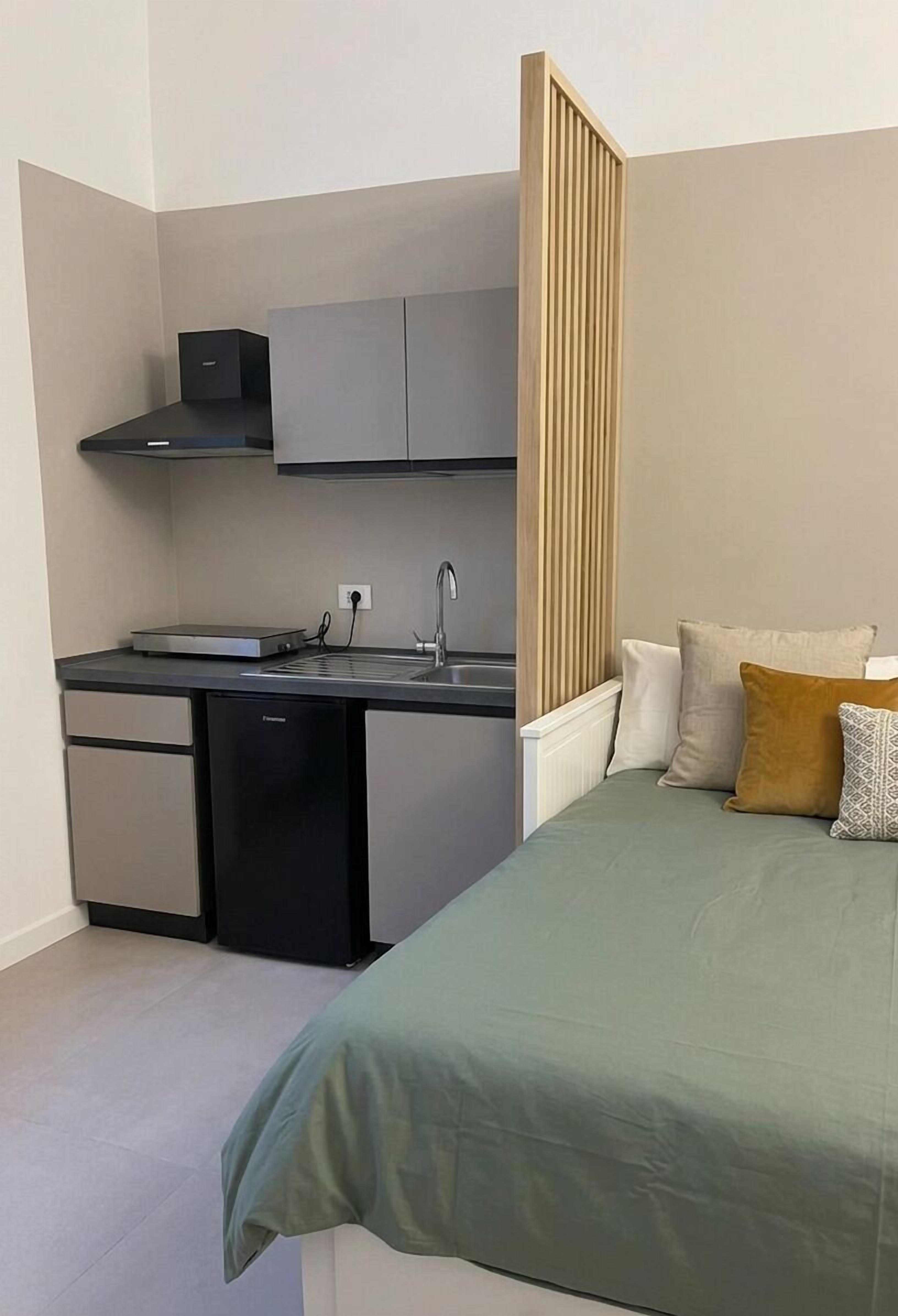 A compact hotel room with a kitchenette, a bed with a green bedspread, and a wooden partition.