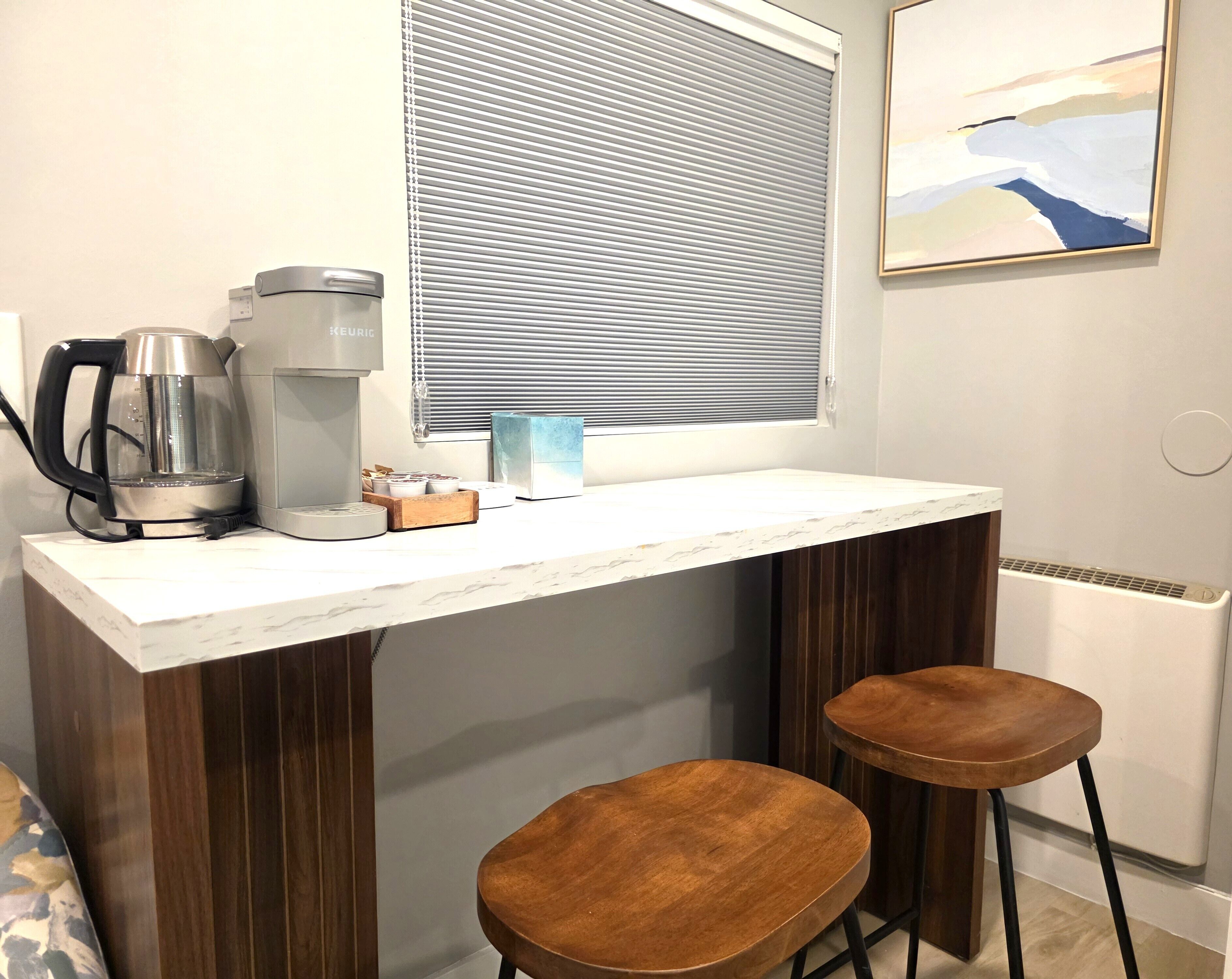A small kitchenette with a coffee maker, a kettle, and a box of tea. There are two wooden stools and a window with blinds.