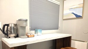 Une petite cuisine équipée d’une machine à café, d’une bouilloire et d’une boîte de sachets de thé. On y trouve deux tabourets en bois et une fenêtre avec des stores.