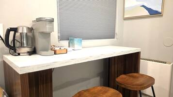 A small kitchenette with a coffee maker, a kettle, and a box of tea. There are two wooden stools and a window with blinds.