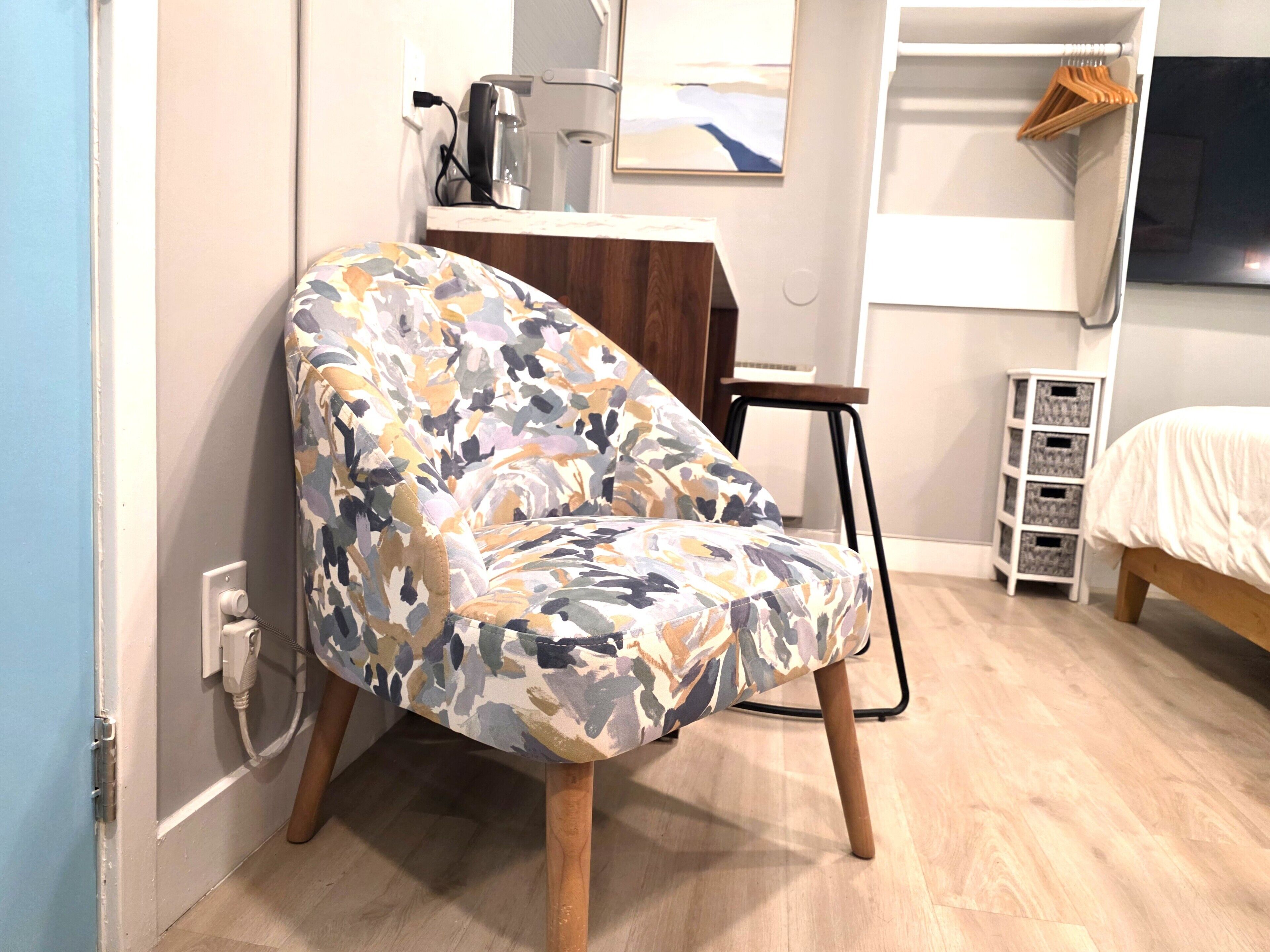Una habitación con un sillón estampado, un escritorio pequeño con un hervidor, una cama con cubrecama blanco y suelo de madera.