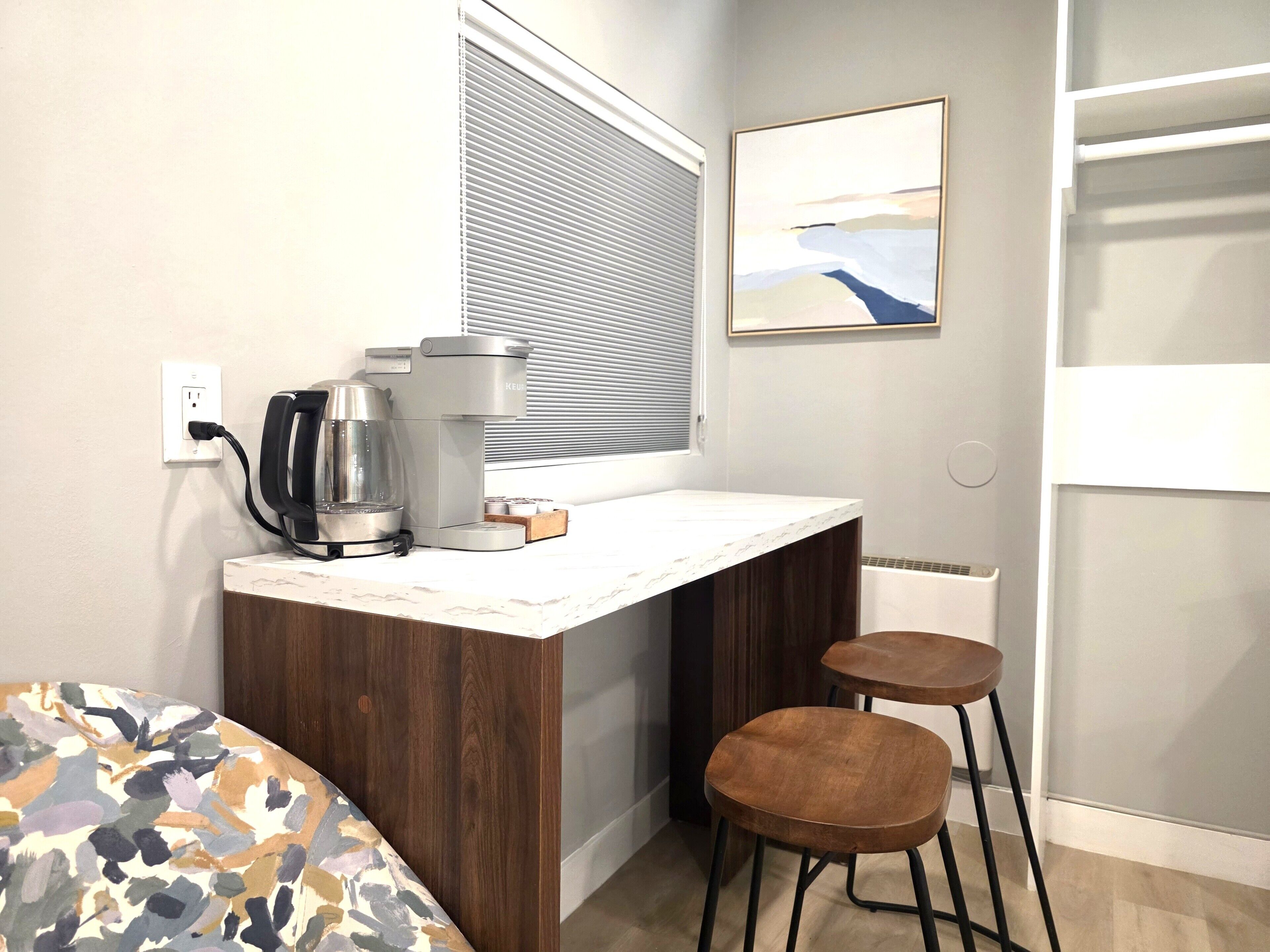 A small kitchenette with a coffee maker, a kettle, and a toaster, a window with blinds, a framed picture on the wall, and a bed with a patterned cover.