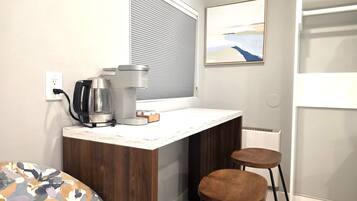 A small kitchenette with a coffee maker, a kettle, and a toaster, a window with blinds, a framed picture on the wall, and a bed with a patterned cover.