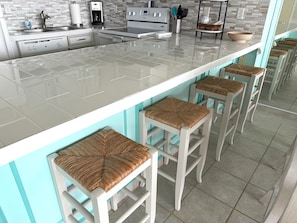 A modern kitchen with a white countertop, stainless steel appliances, and a bar area with white stools. - New Listing! Beachfront 2 Bdrm Condo, Family Friendly. Make great memories here! (South Padre Island)