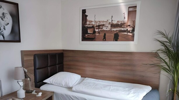 Ein Einzelbett mit Holz-Kopfteil, ein Nachttisch mit Lampe, ein gerahmtes Bild an der Wand und ein Blumentopf.