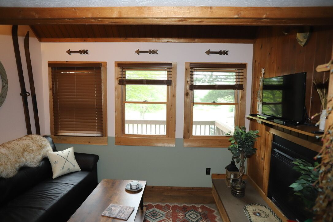 A cozy living room with a black leather sofa, a wooden coffee table, and a television on a wooden stand. There are three windows with wooden frames and blinds, and a fireplace with a plant on the mantel.