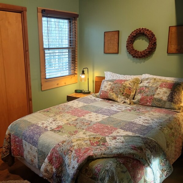 A bedroom with a floral quilt, a wooden headboard, a nightstand with a lamp, a window with blinds, and a wreath on the wall.