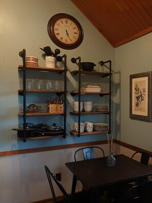 A dining area with a wooden table, chairs, a wall-mounted clock, and shelves with kitchenware. - 1BR Cabin W/ Loft For 2 Great Views Over Resort (Davis)