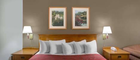 A hotel room with a wooden headboard, a red patterned bedspread, two framed landscape paintings, a bedside table with a lamp, and a telephone.