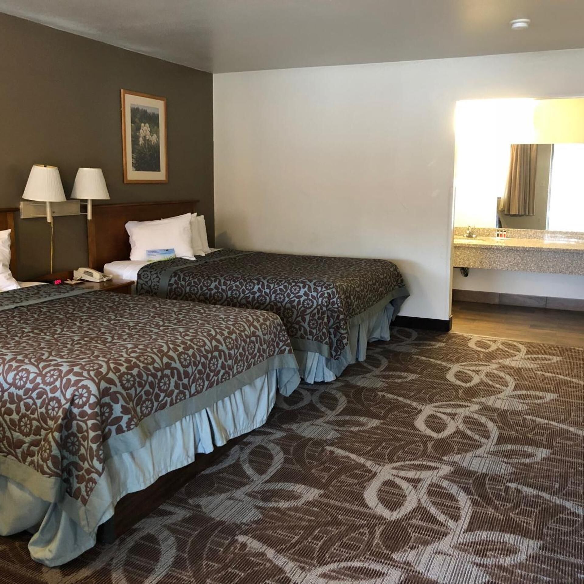 A hotel room with two beds, a nightstand, a lamp, a painting, and a bathroom visible through an open door.