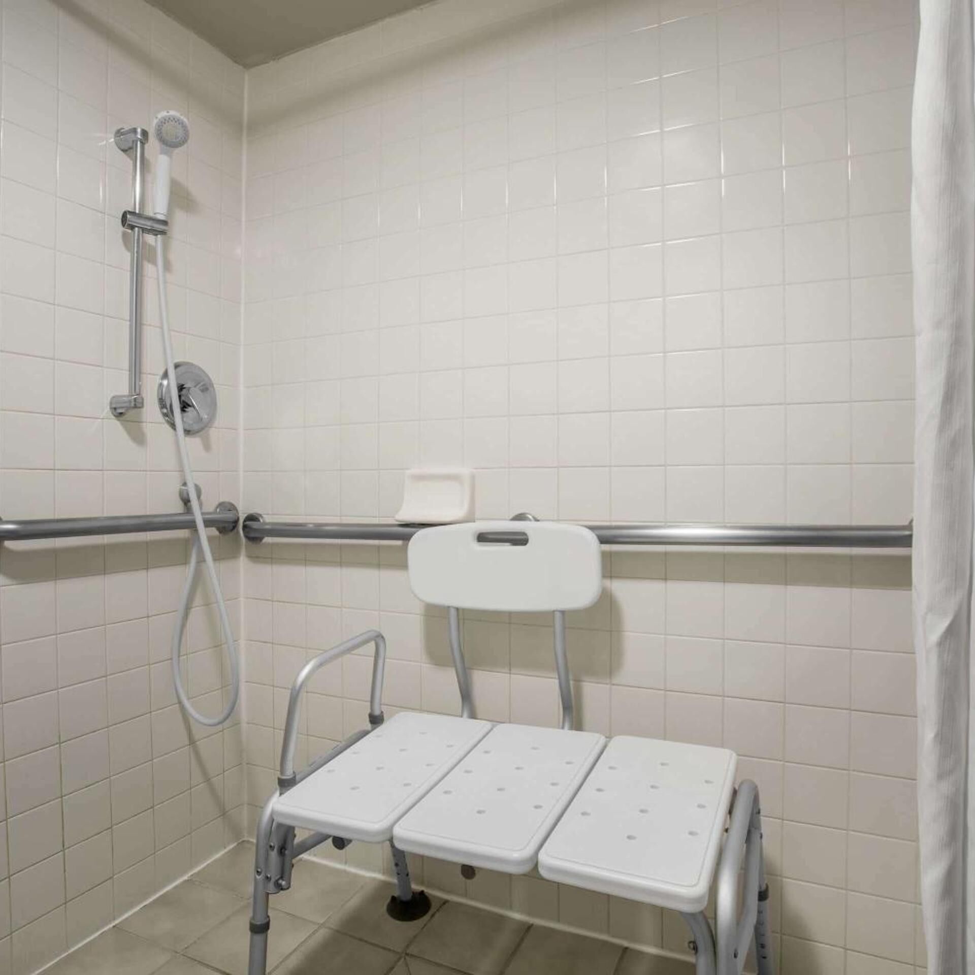 A bathroom with white tiled walls, a shower with a handheld showerhead, a grab bar, and a wheelchair-accessible bench.