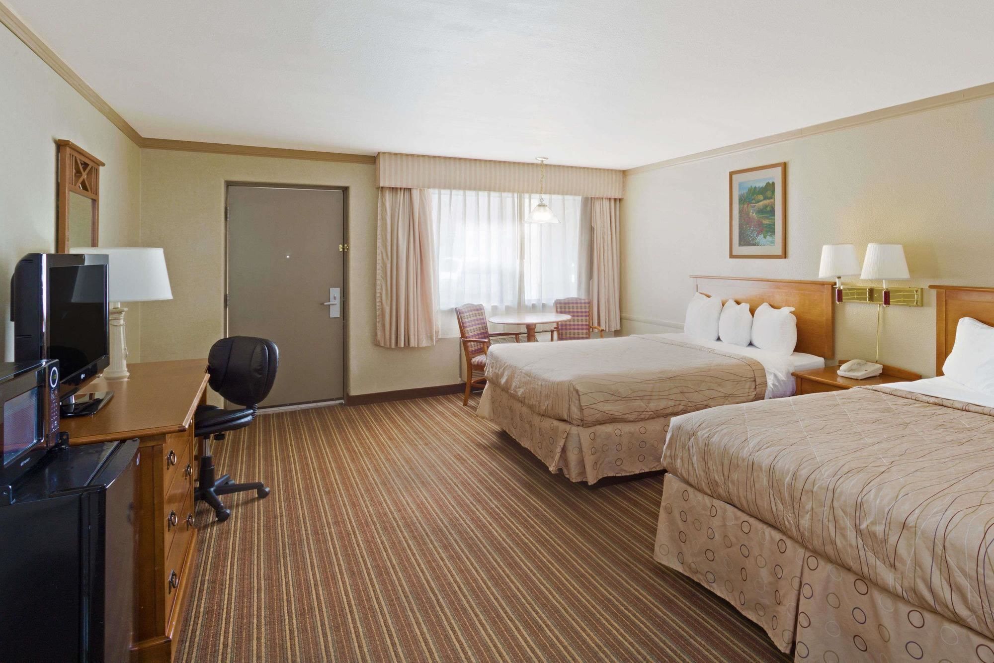 A hotel room with two beds, a desk with a computer, a chair, a small table, and a window with curtains.