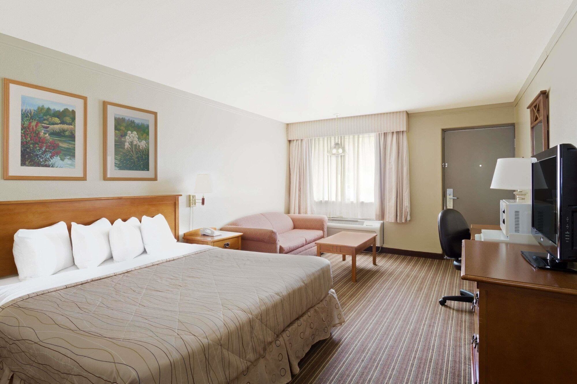 A hotel room with a large bed, a desk with a computer, a chair, a small table, and two framed pictures on the wall.
