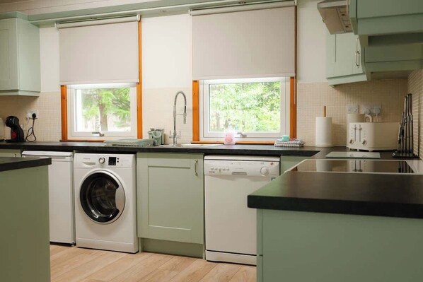 A kitchen with a washing machine, dishwasher, and a window with a view of trees.