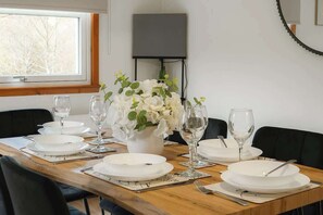 Een eettafel gedekt met witte borden, glazen en bestek, een vaas met witte bloemen, en een aan de muur bevestigde televisie.