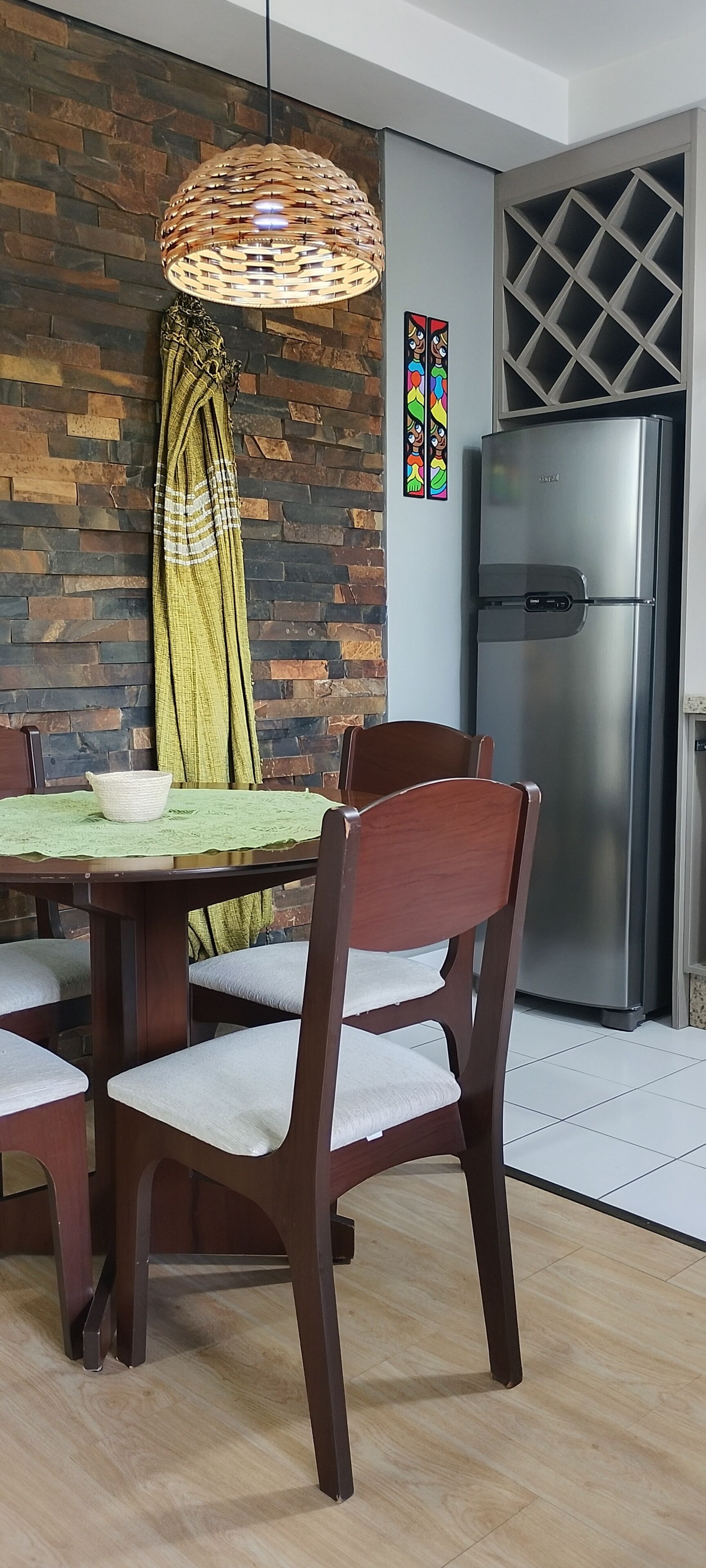 Un coin repas avec une table en bois, des chaises, un luminaire en fibres tressées, un réfrigérateur et un mur en briques.