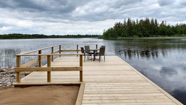 A wooden dock with chairs and a table, overlooking a calm lake with a forested shoreline. - 4 Star Holiday Home in Emmaboda (Emmaboda)