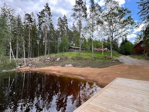 A wooden dock extending over a calm body of water, surrounded by a forest and a dirt path leading to a red cabin. - 4 Star Holiday Home in Emmaboda (Emmaboda)