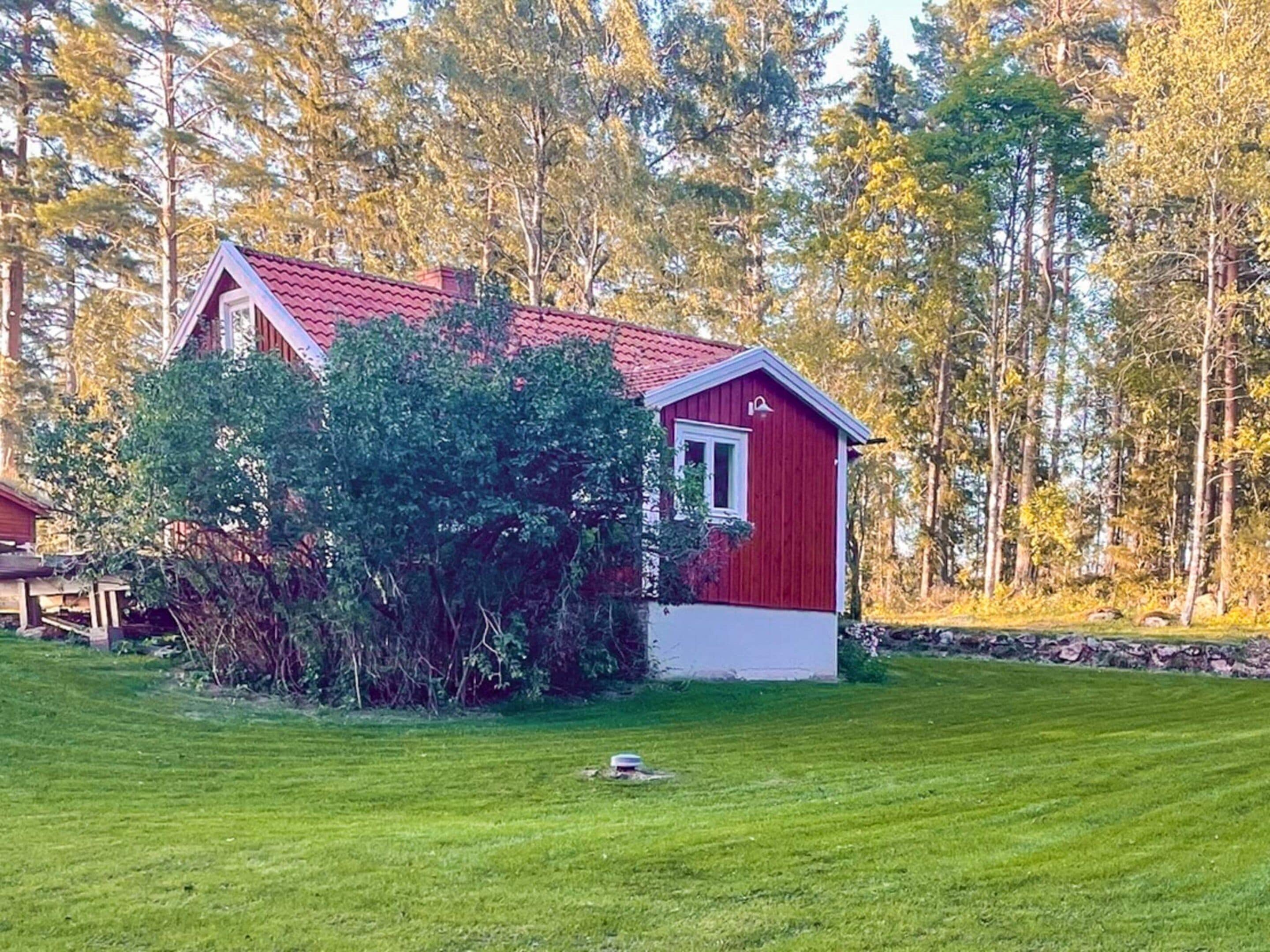 A red wooden house with a white trim, surrounded by a well-maintained lawn and dense trees in the background.