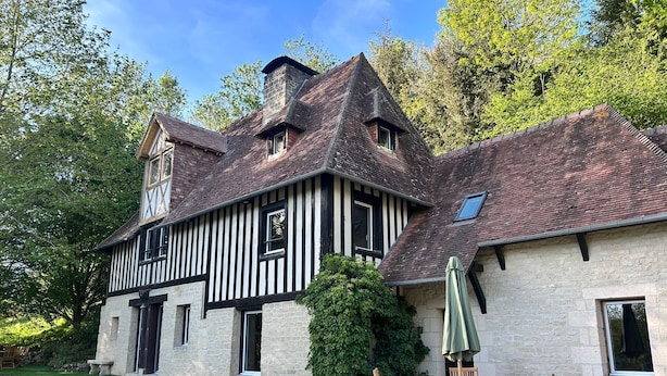 Um edifício tradicional em estilo de enxaimel, com chaminé de pedra, uma pequena janela e um pátio de brita com mesa e cadeiras de madeira.