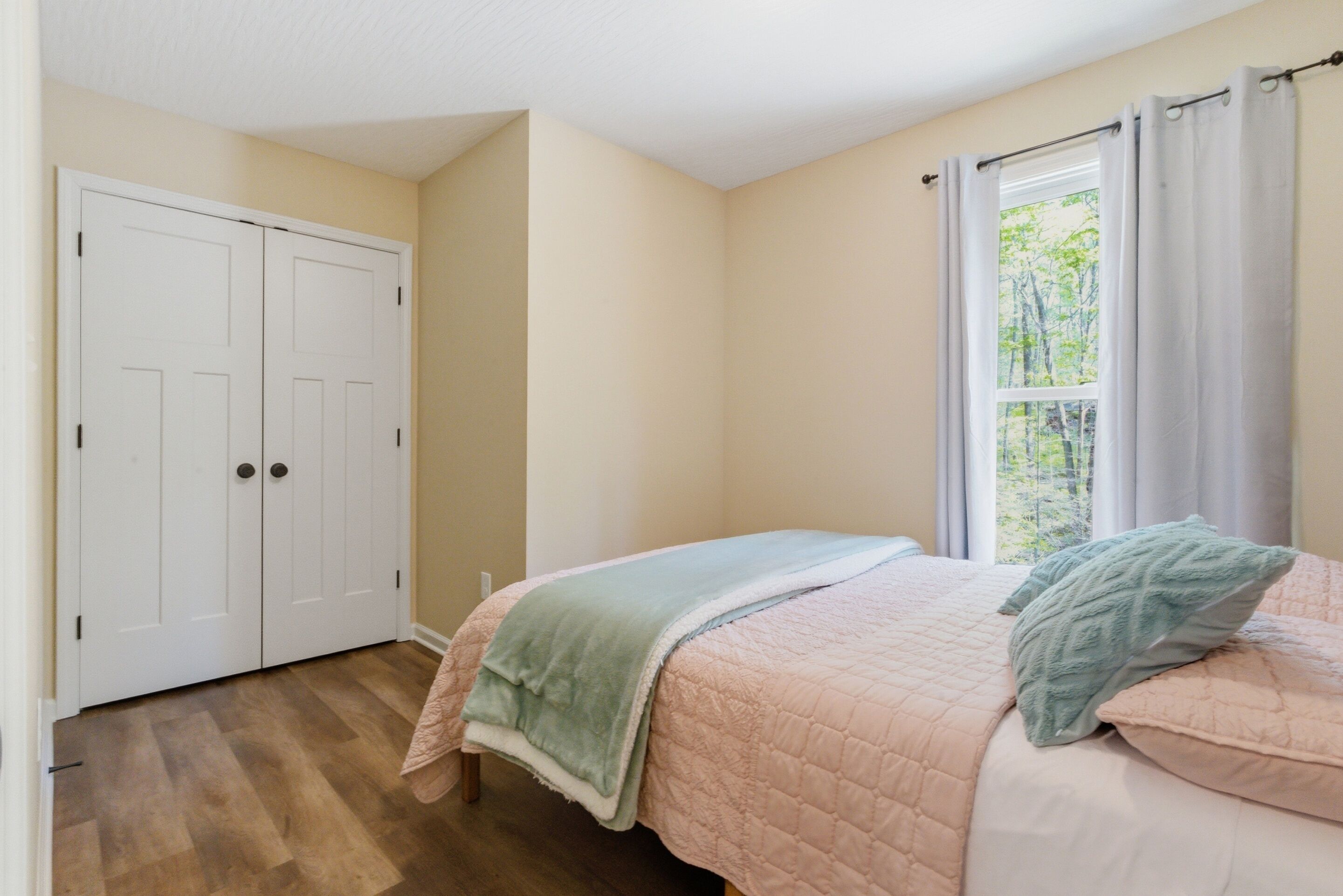 Un dormitorio con cama, ventana con cortinas, puerta y piso de madera.