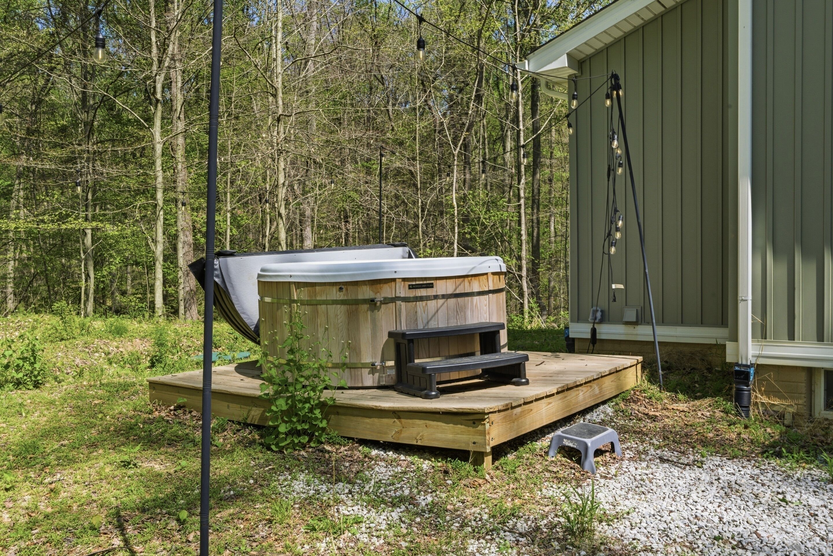 Un bain à remous doté d’une terrasse en bois et d’un petit tabouret, entouré d’une zone boisée.